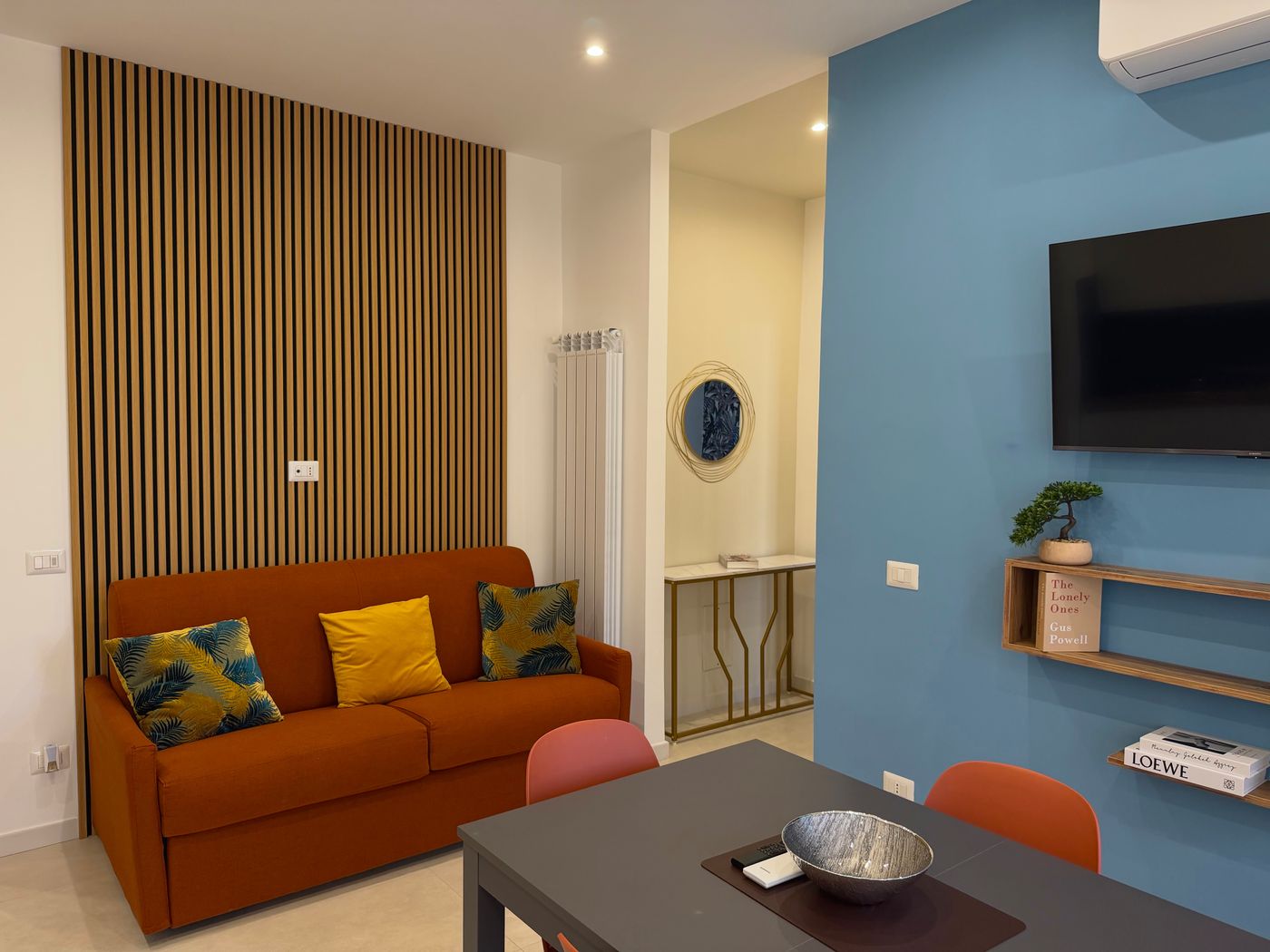 Divano letto arancione, tavolo da pranzo e parete azzurra con la TV