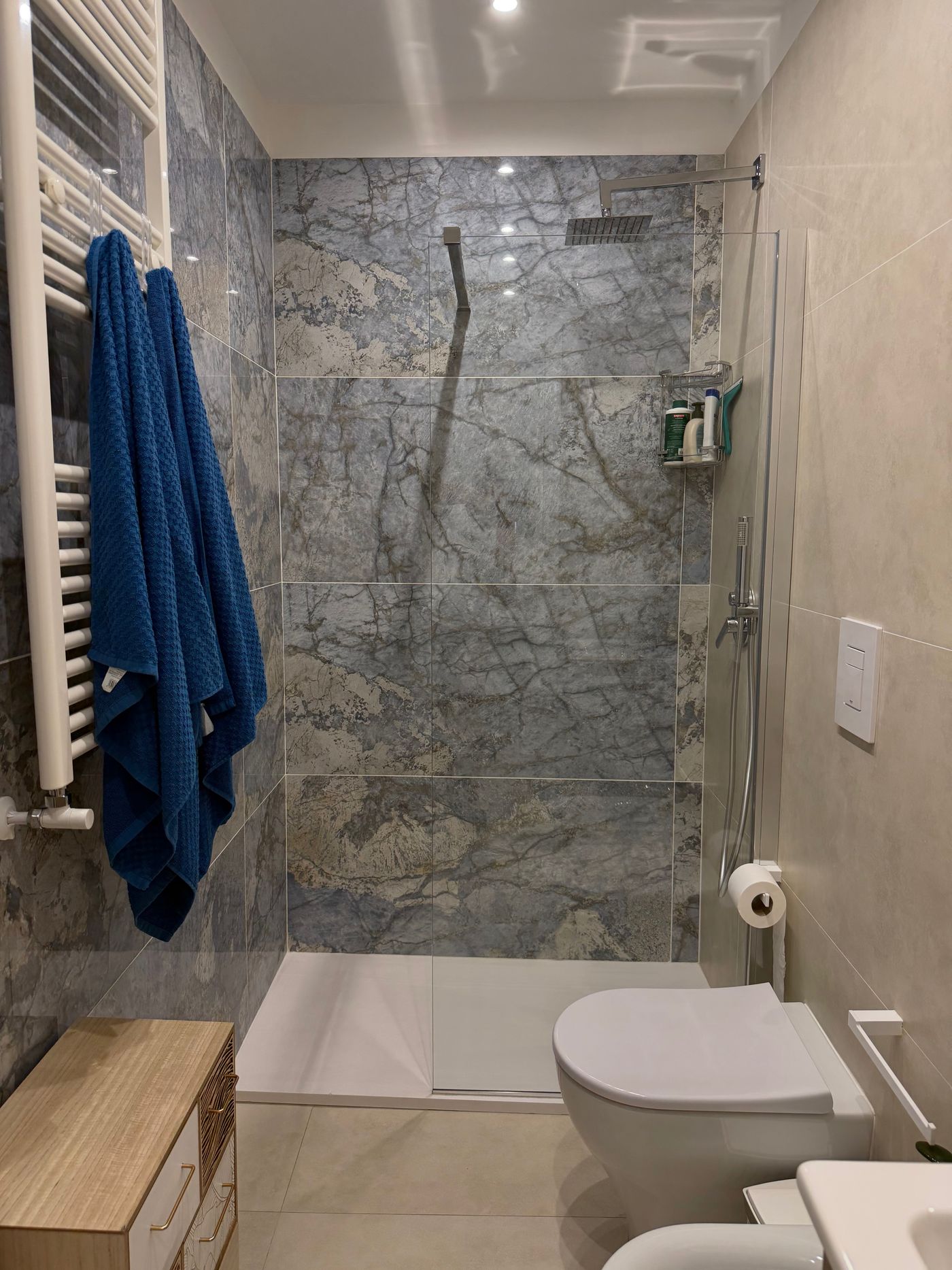Bagno con doccia rivestita in marmo grigio-azzurro e asciugamani blu