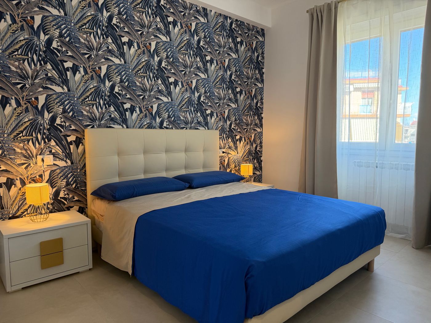 Camera matrimoniale con carta da parati a foglie blu, testiera imbottita e copriletto blu