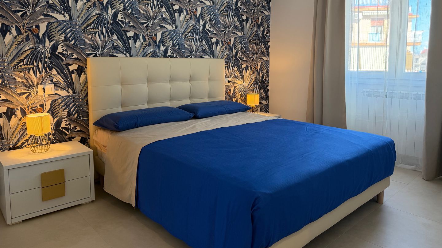 La camera matrimoniale di Monì Boutique, con carta da parati a foglie blu, testiera imbottita e copriletto blu