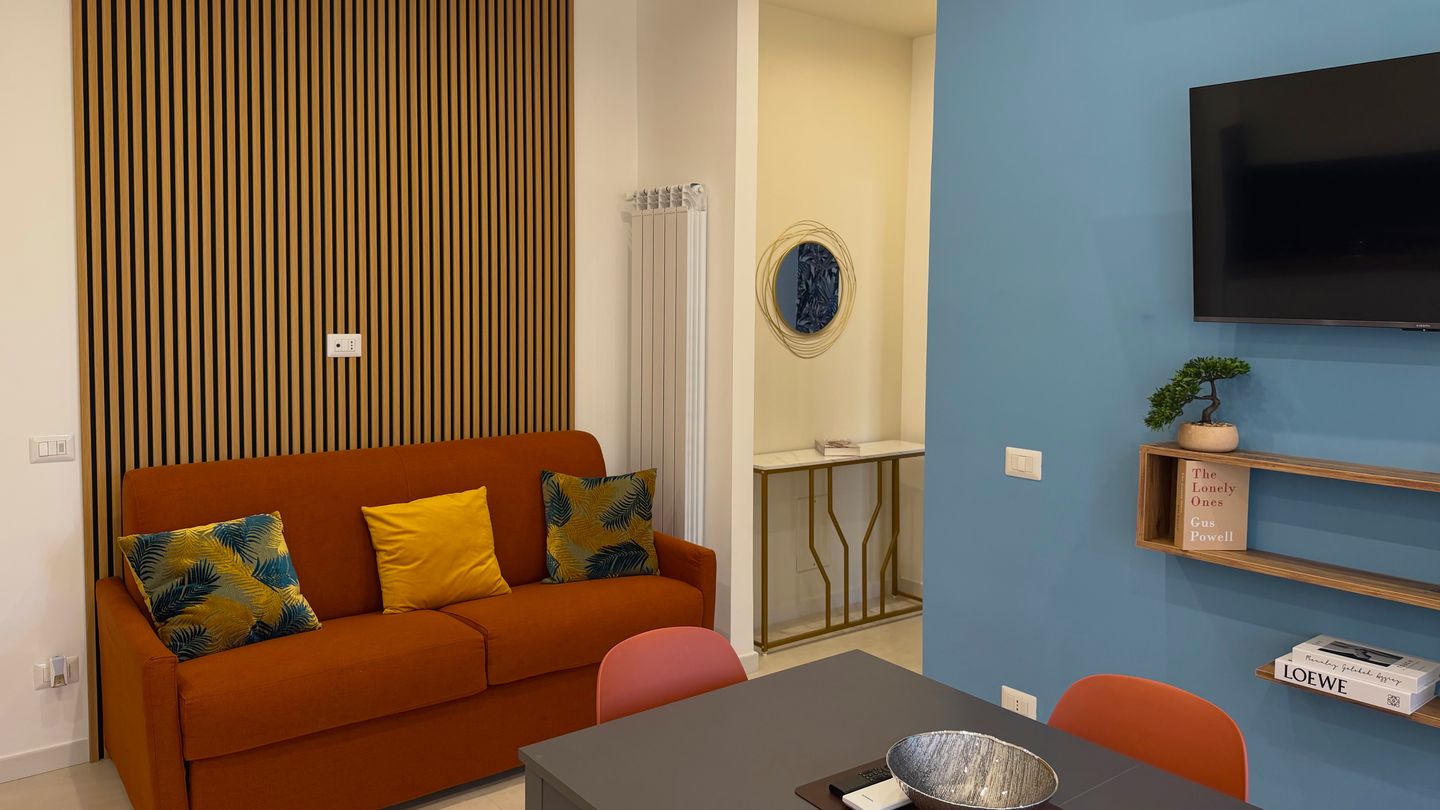 Divano letto arancione, tavolo da pranzo e parete azzurra con la TV